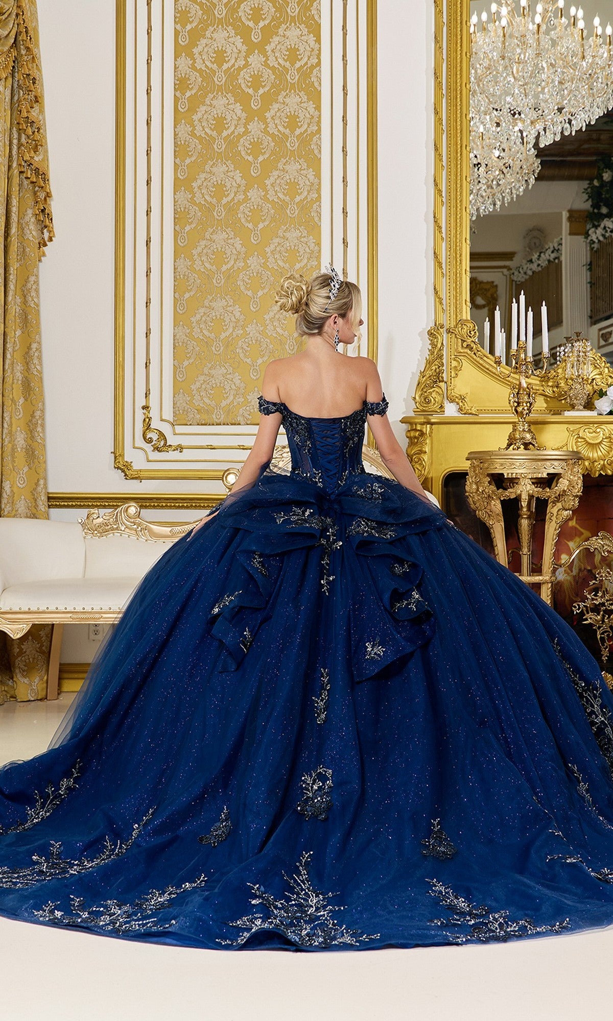 A woman wearing the Quinceanera Dress 5102 By Dancing Queen—navy blue with silver embellishments and lace embroidery—stands facing away in an ornate, gold-accented room.
